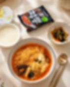 A cozy meal setup featuring a bowl of Pulmuone Roasting Jjamppong topped with egg and seafood, served alongside the Pulmuone Roasting Jjamppong package, a bowl of porridge, a small side dish, and spoon and chopsticks on a light countertop.