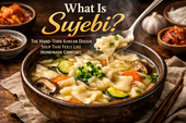 Steaming bowl of Korean sujebi soup in a rustic brown bowl, topped with scallions, zucchini, carrots, and mushrooms, with a spoon lifting a glossy hand-torn dough piece above the broth and headline text asking “What Is Sujebi?”