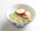 A patterned bowl of pale noodles is topped with cucumber strips, bean sprouts, red sauce, sesame seeds, and half a boiled egg on a light background.