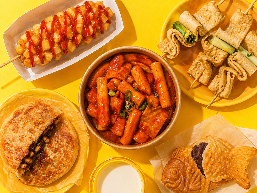 A bright overhead spread of Korean street food on a yellow background shows tteokbokki in the center, a potato-crusted Korean hot dog with ketchup at top left, fish cake skewers at top right, hotteok at bottom left, bungeoppang at bottom right, and a small cup of milk below.