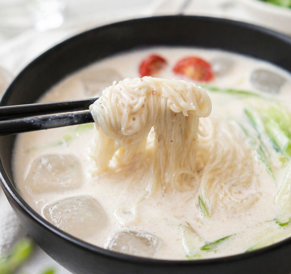 Black chopsticks lift a bundle of thin noodles from a black bowl of cold creamy noodle soup with ice, shredded cucumber, and cherry tomatoes, set in the same bright, clean kitchen setting.