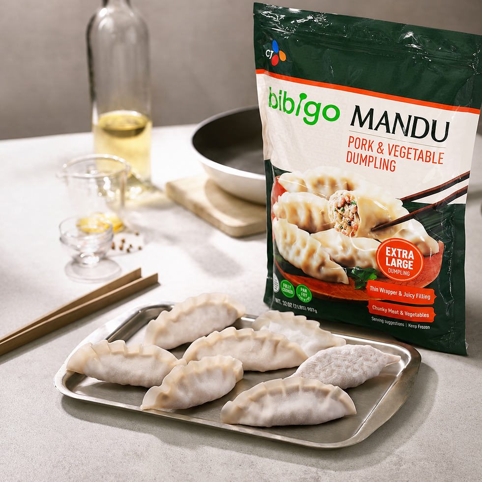 Product lifestyle photo showing CJ Bibigo pork and vegetable extra large dumpling packaging on a kitchen counter beside frozen dumplings arranged on a metal tray, with cooking tools and oil softly blurred in the background.