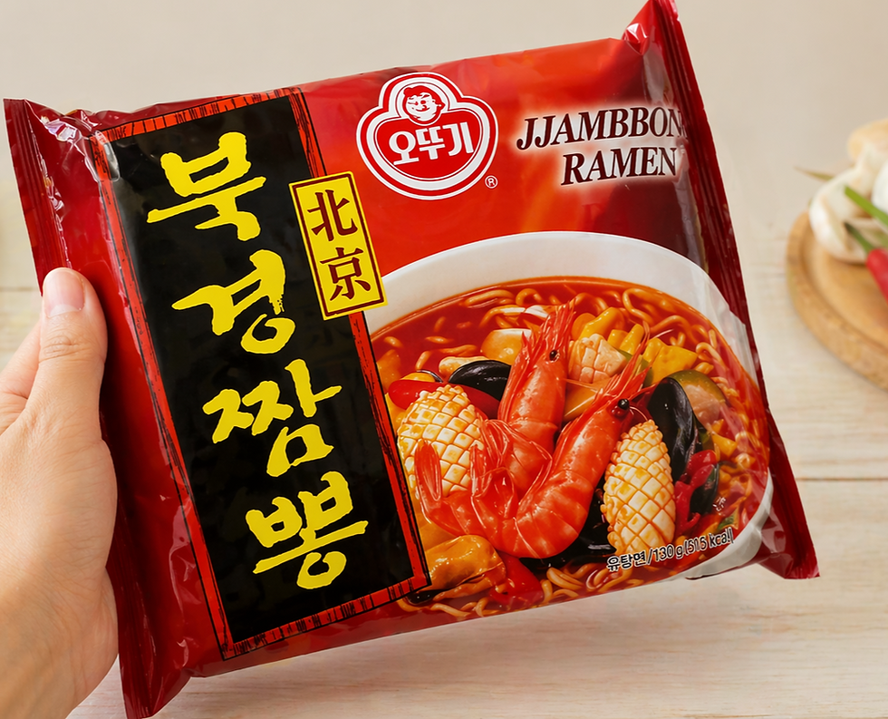 A hand holds a single red package of Ottogi Jjambbong Ramen in a bright kitchen-style setting, with the seafood noodle illustration visible on the front and garlic and red chili peppers in the background.