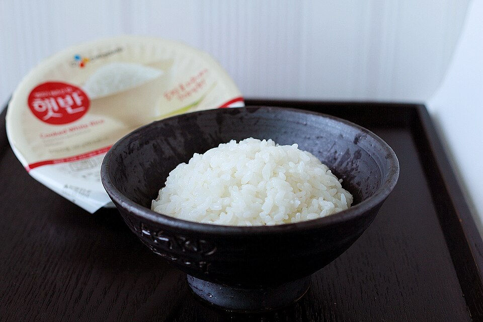 A black bowl filled with steamed white rice on a tray, with instant cooked rice packaging in the background.