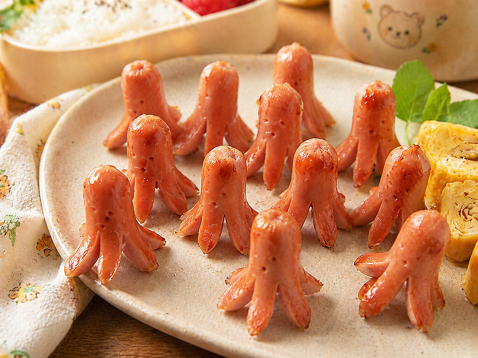 Octopus-shaped Korean fish sausages arranged on a ceramic plate, lightly grilled and glossy, served with rice, rolled omelet, and lunchbox sides in a warm Korean kitchen table setting.