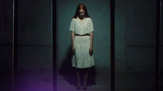 Docufiction “Il Muro”: ragazza con capelli lunghi e vestito bianco sta in piedi in un corridoio buio, atmosfera inquietante.