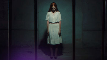 Docufiction “Il Muro”: ragazza con capelli lunghi e vestito bianco sta in piedi in un corridoio buio, atmosfera inquietante.