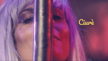 Scena del film Ciurè: donna con parrucca biondo platino e trucco glitterato, primo piano con luci rosa e viola.