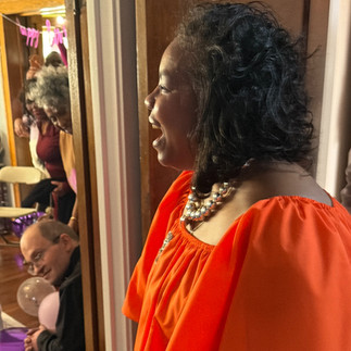Woman in orange dress reacting with surprise and joy after entering her 60th birthday celebration surrounded by guests and purple decorations.