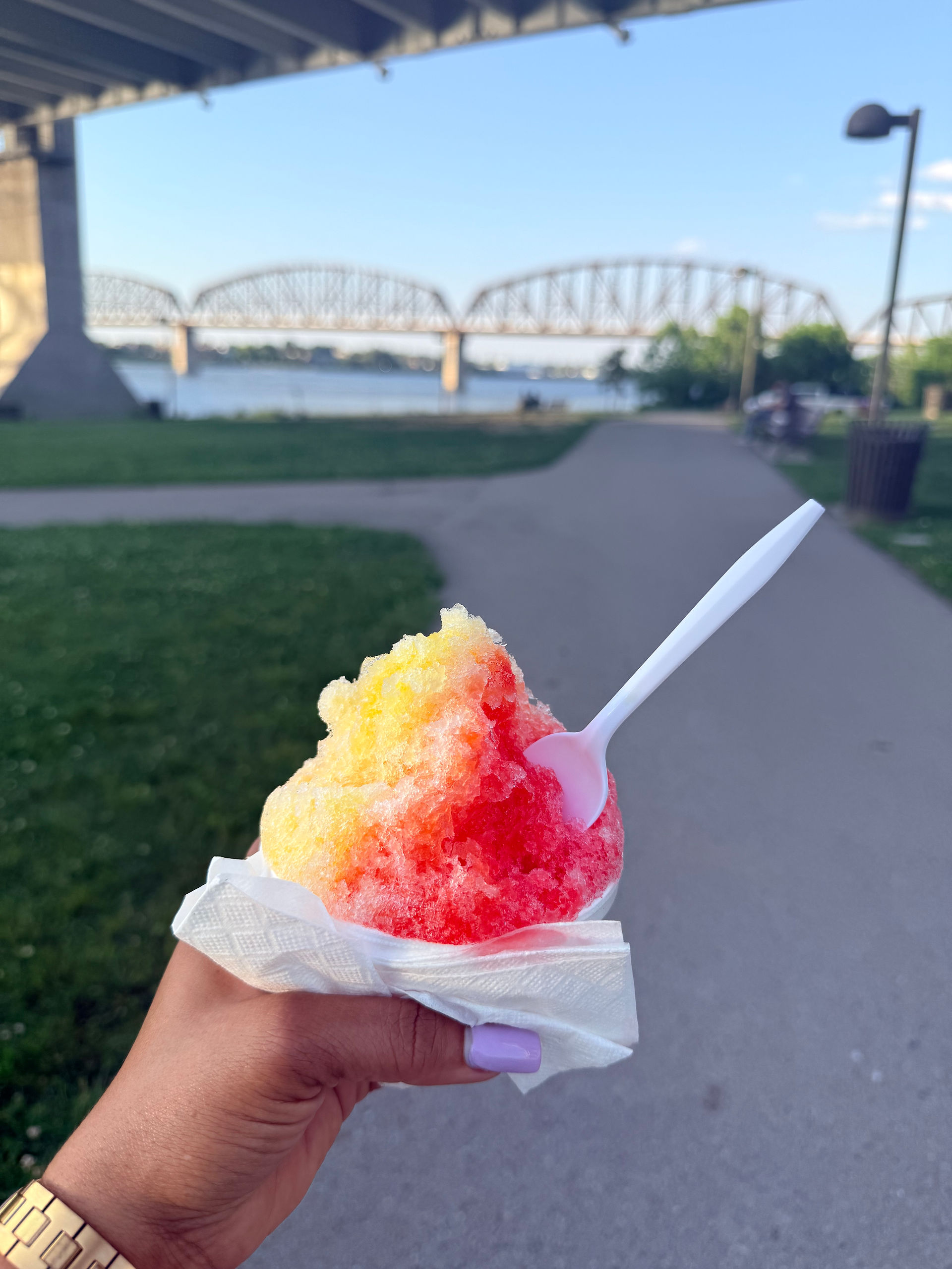 Hand holding a colorful Kona Ice shaved ice treat with the Big Four Bridge and Ohio River in the background in Louisville Kentucky