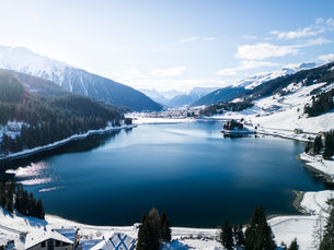 Winterliche Schweizer Alpenlandschaft mit verschneiten Bergen, tiefblauem Bergsee und Ortschaft im Hintergrund.
