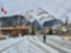 Snowy street in a mountain town, with a person cycling. Canadian flags wave near rustic shops. Snowy peaks and cloudy sky in the background.