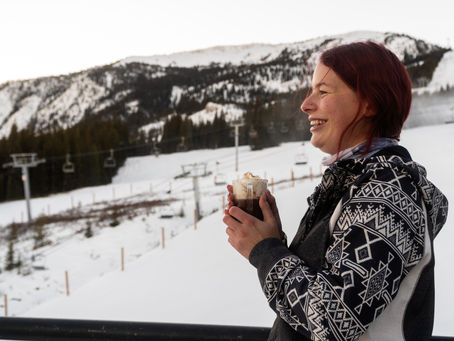 Woman with red hair, smiling and holding a hot drink, stands in snowy mountain setting with ski lifts and pine trees in the background.