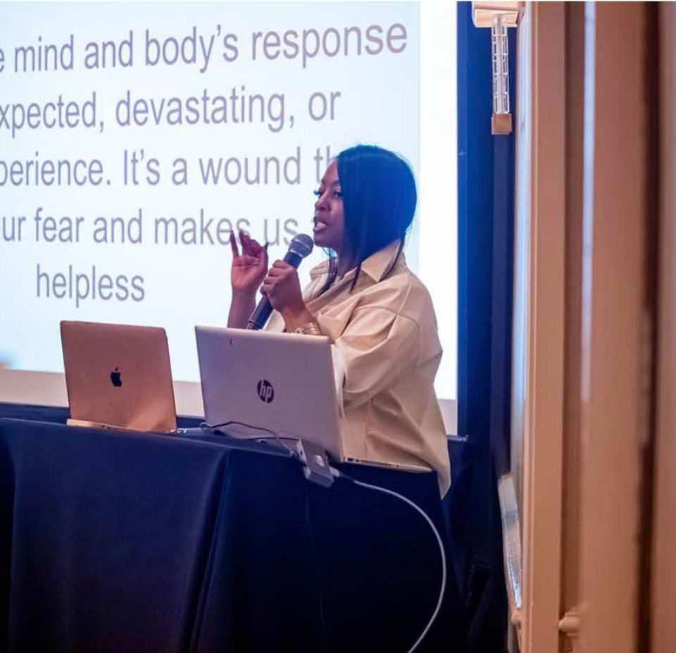 Clara Throckmorton delivering a presentation about trauma, speaking into a microphone with slides projected behind her