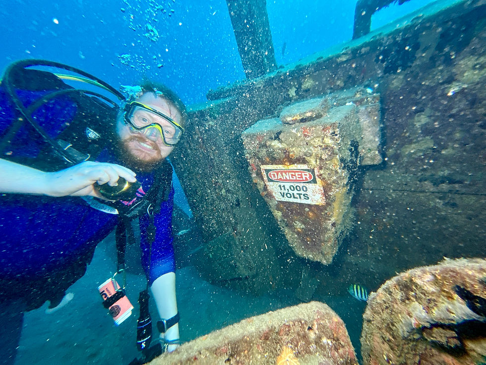 Scuba diver poses with a Big smile underwater near a rusted shipwreck with a "DANGER 11,000 VOLTS" sign. Blue water, a small fish, and a relaxed, humorous mood.
