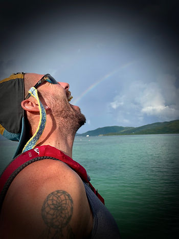Man smiling at sky, wearing sunglasses and cap. Visible tattoo on shoulder. Background: ocean, mountains, rainbow, and cloudy sky. Man positioned with mouth open to make it look as if the rainbow is coming out of his mouth.