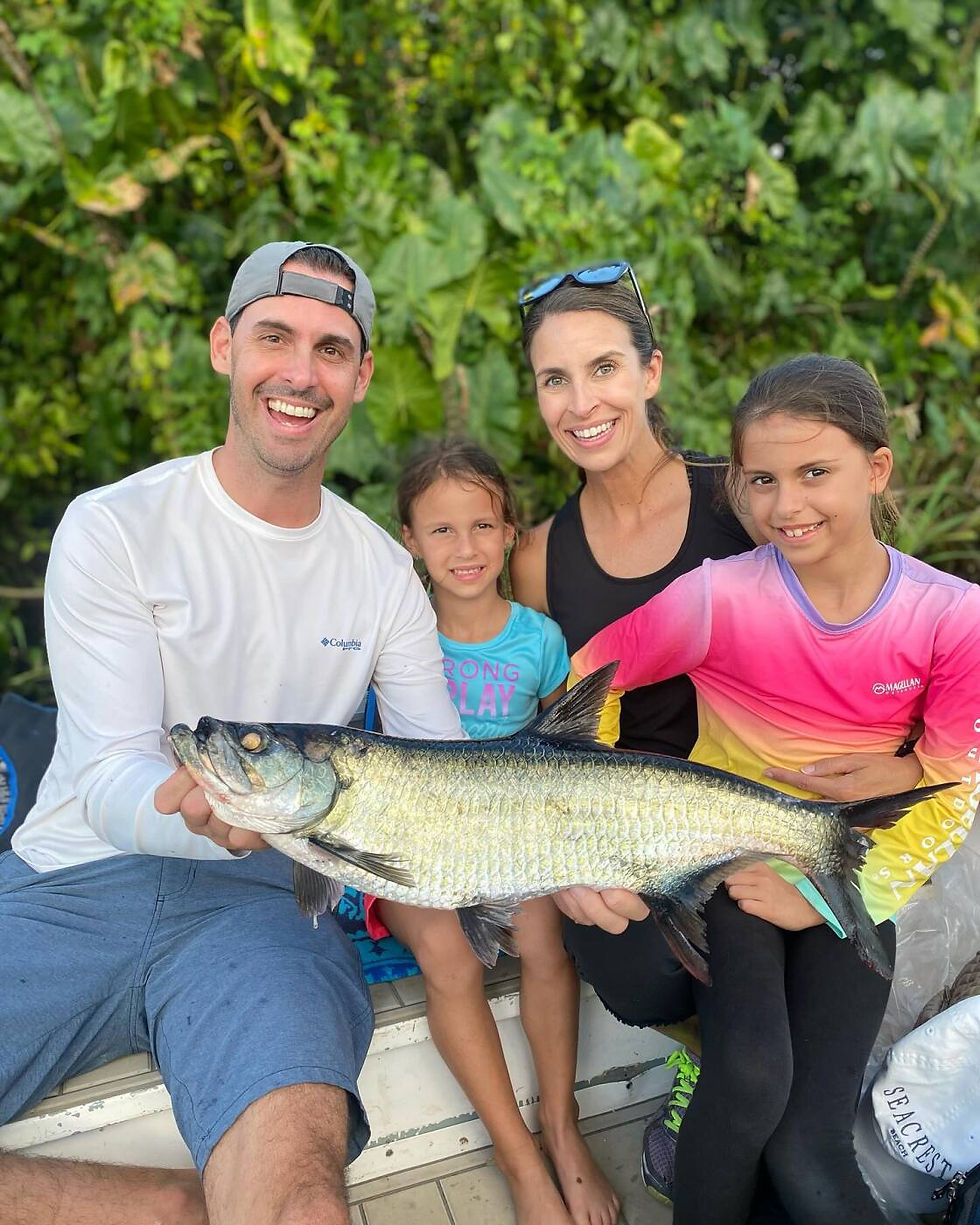 Family of four smiles holding a large fish on a boat. Lush green foliage in the background. Bright shirts add a cheerful vibe.