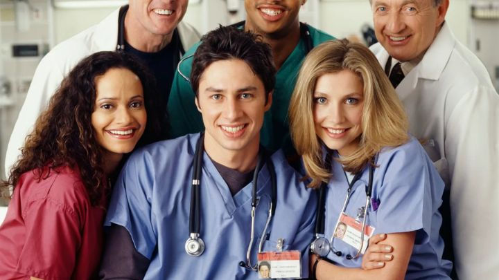 Carla, J.D., and Elliot smile in the foreground while Dr. Cox, Turk, and Kelso stand behind them in this classic Scrubs cast group photo.