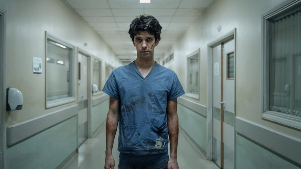 Ben Whishaw as Adam Kay in This Is Going to Hurt, standing in a hospital hallway wearing blood-stained scrubs and looking exhausted.