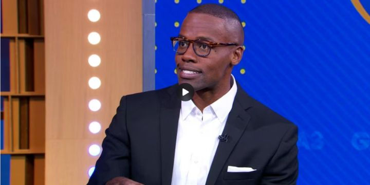 A man in a black suit and glasses speaking during an interview on a set, with a video play button visible in the corner.