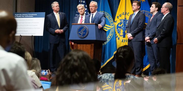 Group of officials standing on a stage at a press conference, with one man speaking at a podium featuring the U.S. Department of Health and Human Services seal, and others standing beside him in front of blue and yellow HHS and U.S. Public Health Service flags.