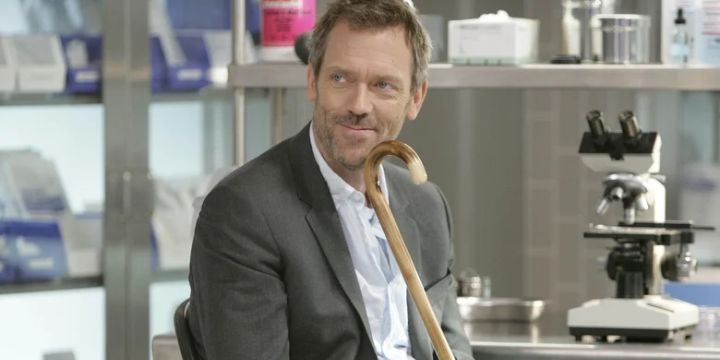 Hugh Laurie as Dr. House, holding his cane and smiling subtly in a medical lab setting.