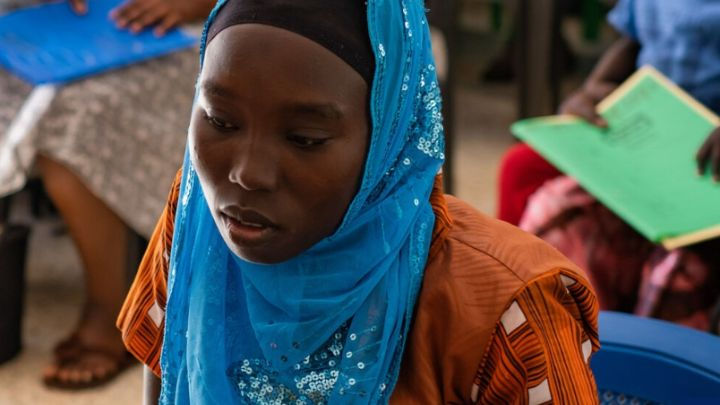 Young woman in a blue headscarf and orange patterned dress, looking down thoughtfully during a meeting or class.
