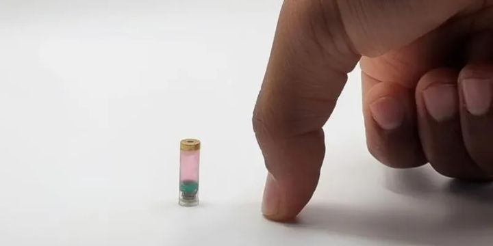 A human finger next to a tiny, pink and teal ingestible bioprinter capsule, illustrating its remarkably small size.