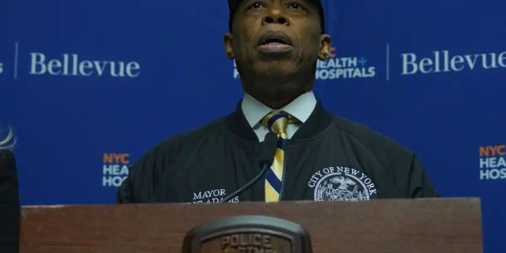 NYC Mayor Eric Adams speaks at a press conference in front of a blue backdrop with "Bellevue" visible.