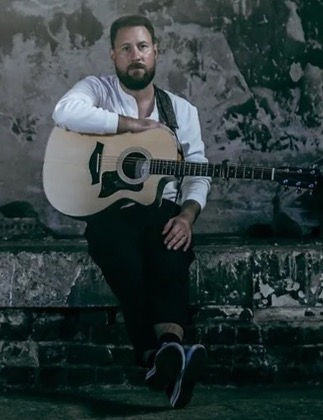 Nick Edwards sits on a concrete step with his guitar looking at the camera.