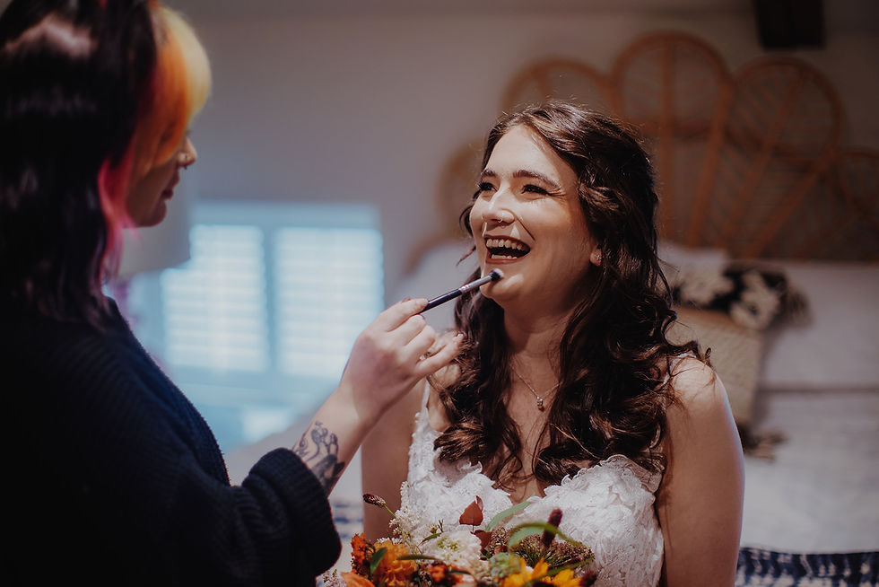 A bride laughs as a makeup artist applies makeup with a brush. The setting is cozy with a bright window, wicker decor, and floral details.