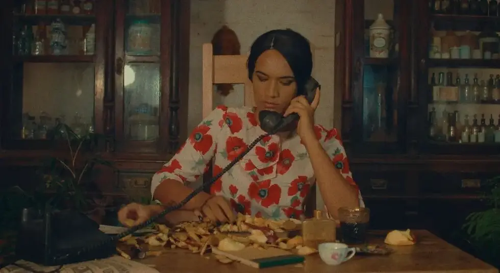 Mulher sentada em uma cozinha vintage, usando um vestido floral colorido, segura o telefone enquanto descasca vegetais, cercada por xícaras e potes, em um ambiente antigo e acolhedor.