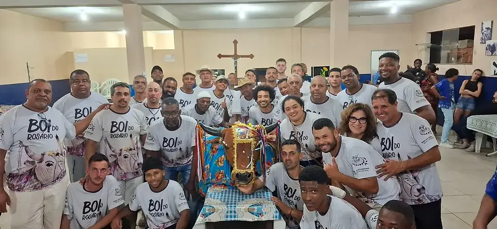 Grupo de homens e mulheres com camisetas BOI do Rosário posa em salão, cercando uma cabeça de boi decorada; cruz ao fundo.