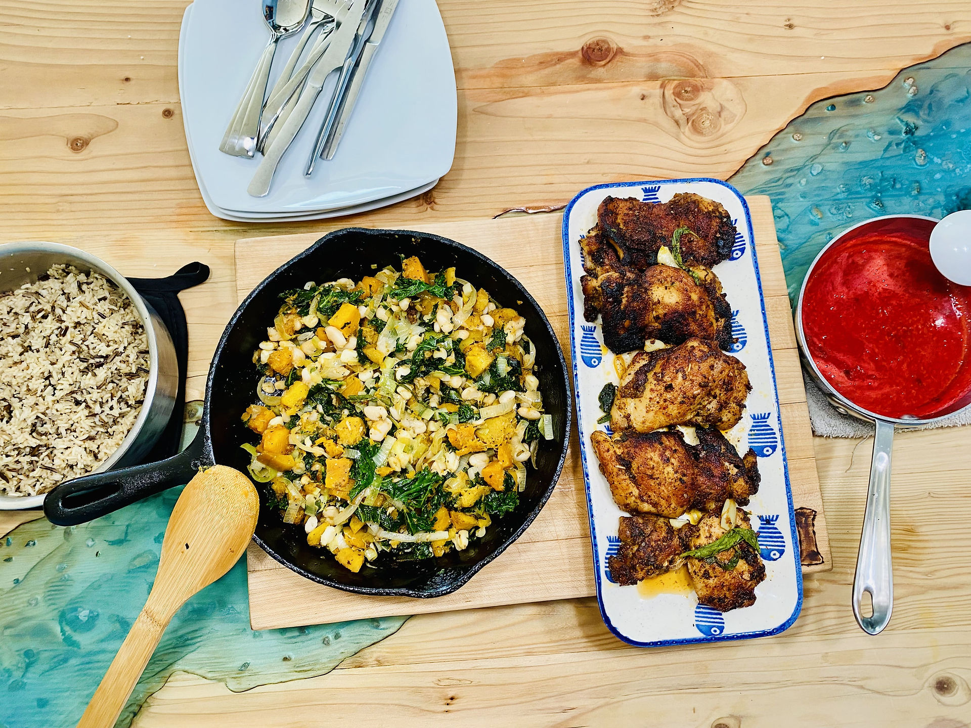 From the left: Wild Rice in a stainless steel pot, Roasted Root Vegetables in a cast iron and Pan Seared Chicken Thigh on a ceramic rectangular plate with blue fish prints, and raspberry sauce in a small stainless steel pot, all on the dining table.  