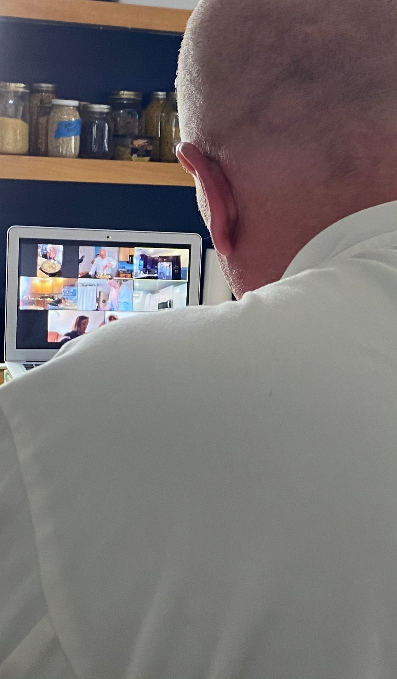 Chef Aric's shoulder in a white chef jacket, with zoom members in the background cooking together.