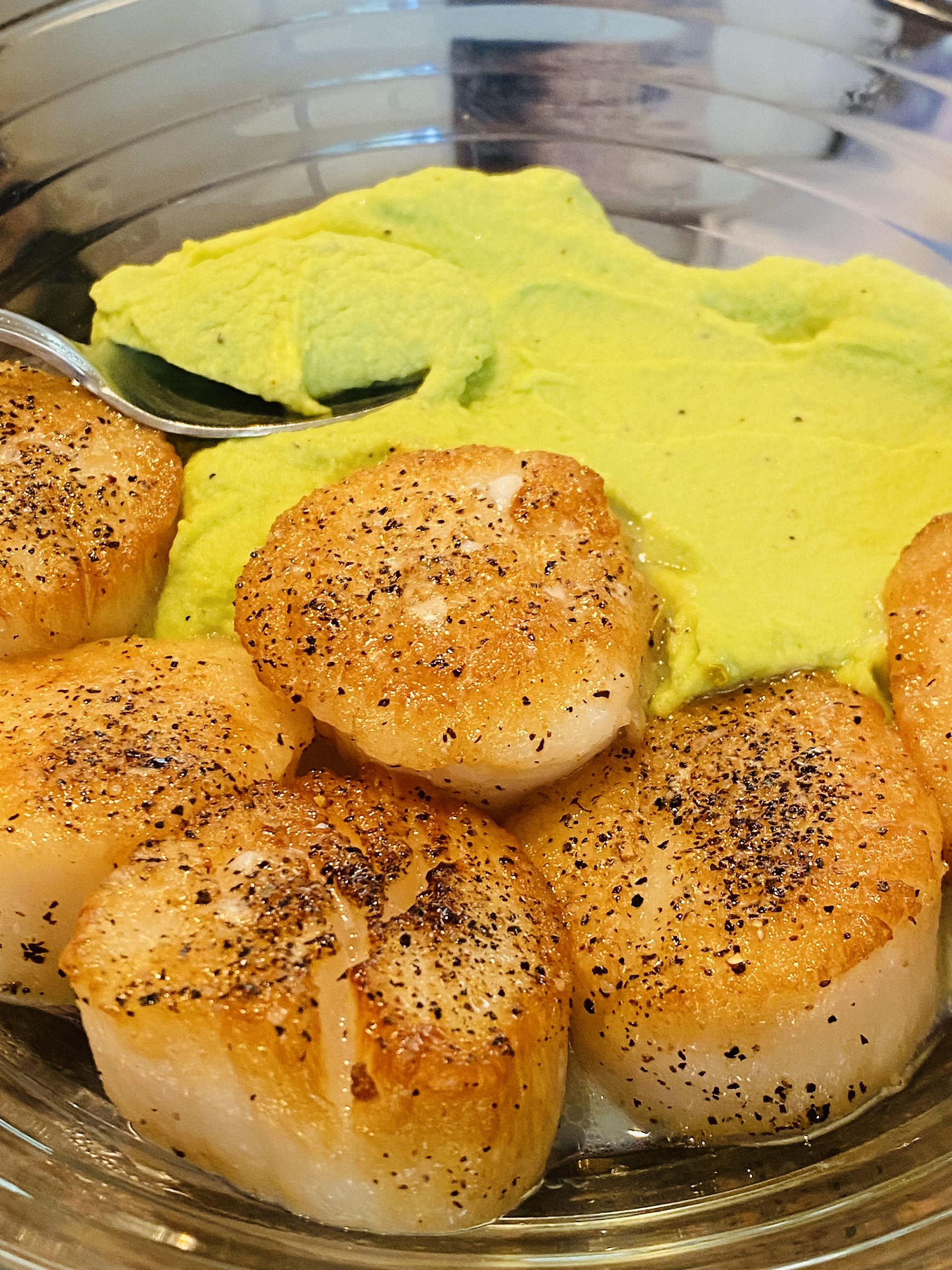 Asian Fusion Sea Scallops beautifully seared with salt and pepper on edamame puree on the clear glass plate with a spoon