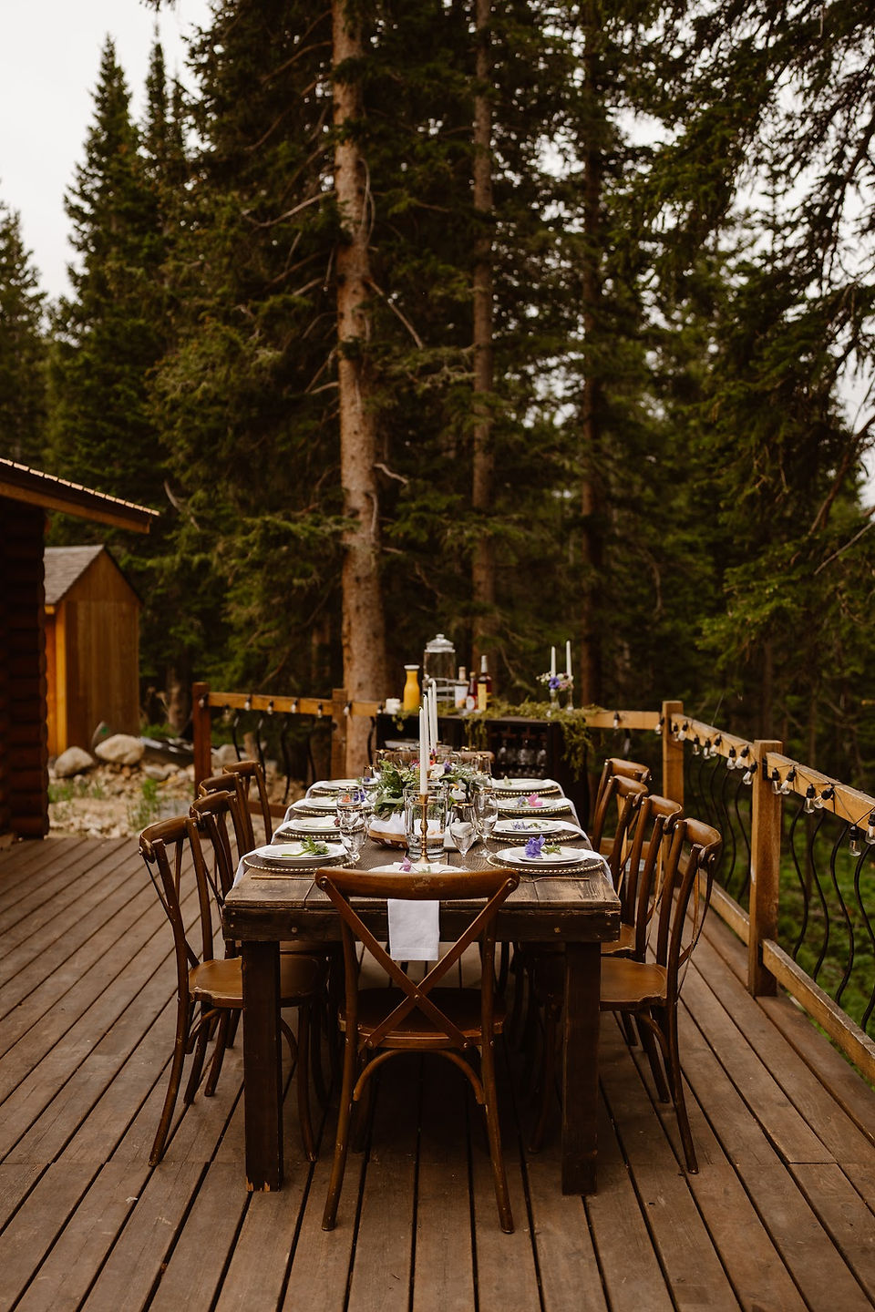 Outside on a wooden balcony with wooden railing and large green trees in the background sits a rectangular wooden table with 10 chairs. White plates and silverwear are in front of each chair on the table and white candles are in the center of the table.