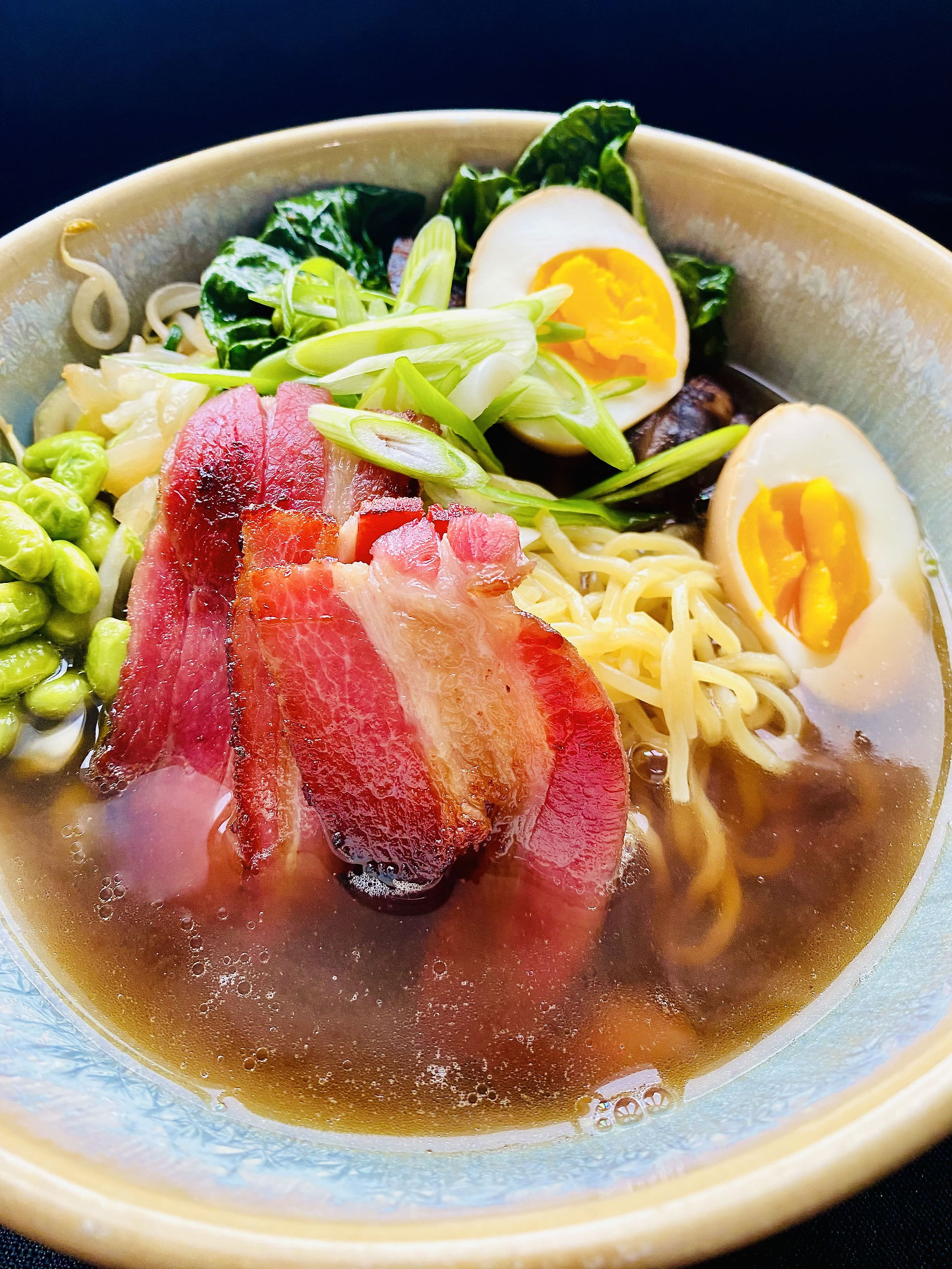 A beautiful photo of a ramen noodles with edamame, spinach, boiled egg cut in half, and homemed bacon in the middle of the ramen, julienne scallions on top as a garnish.