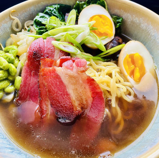 A beautiful photo of a ramen noodles with edamame, spinach, boiled egg cut in half, and homemed bacon in the middle of the ramen, julienne scallions on top as a garnish.