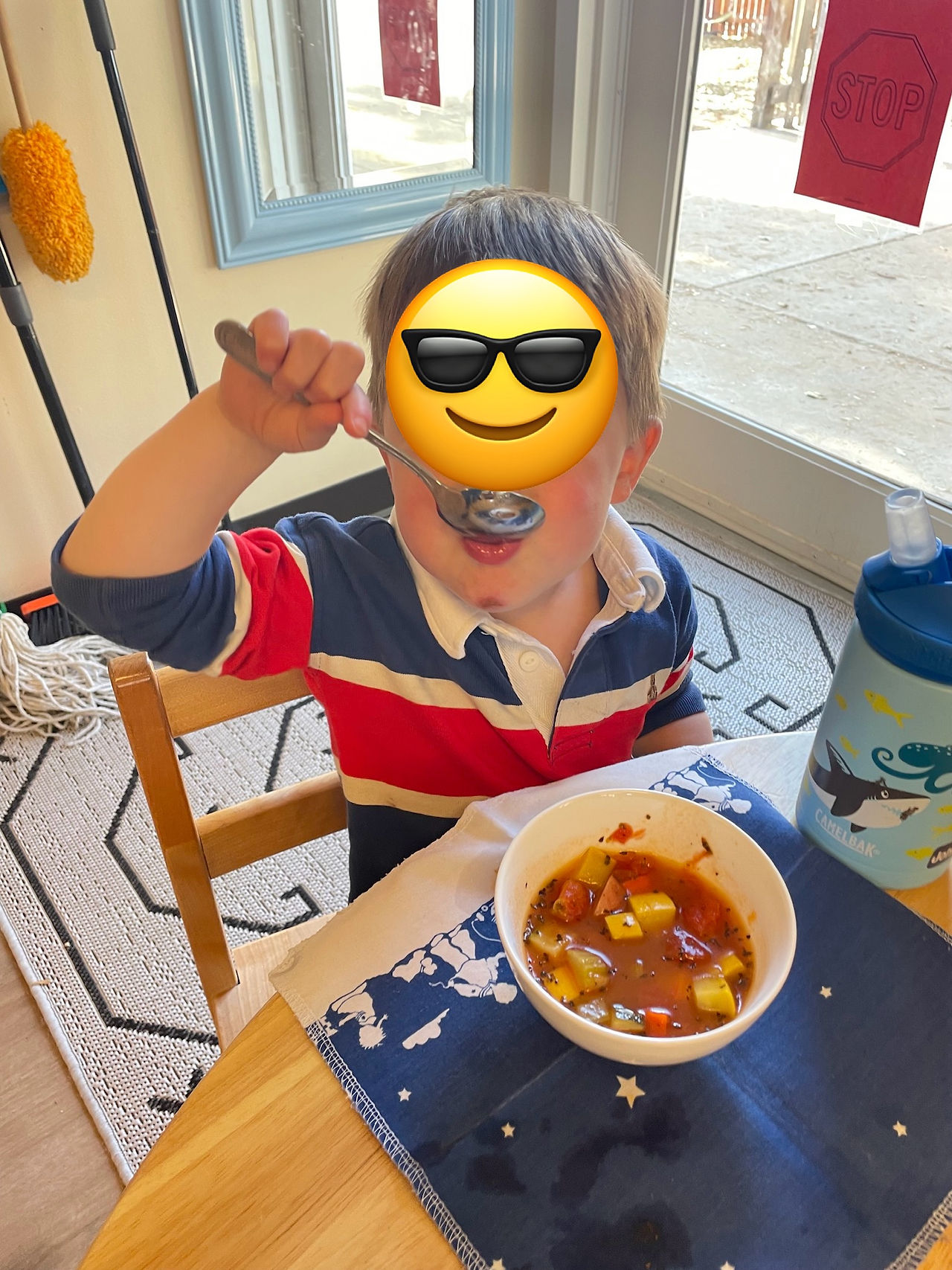 A 3 year old boy (face hidden by a sticker) putting a spoon in his mouth, enjoying his vegetable soup