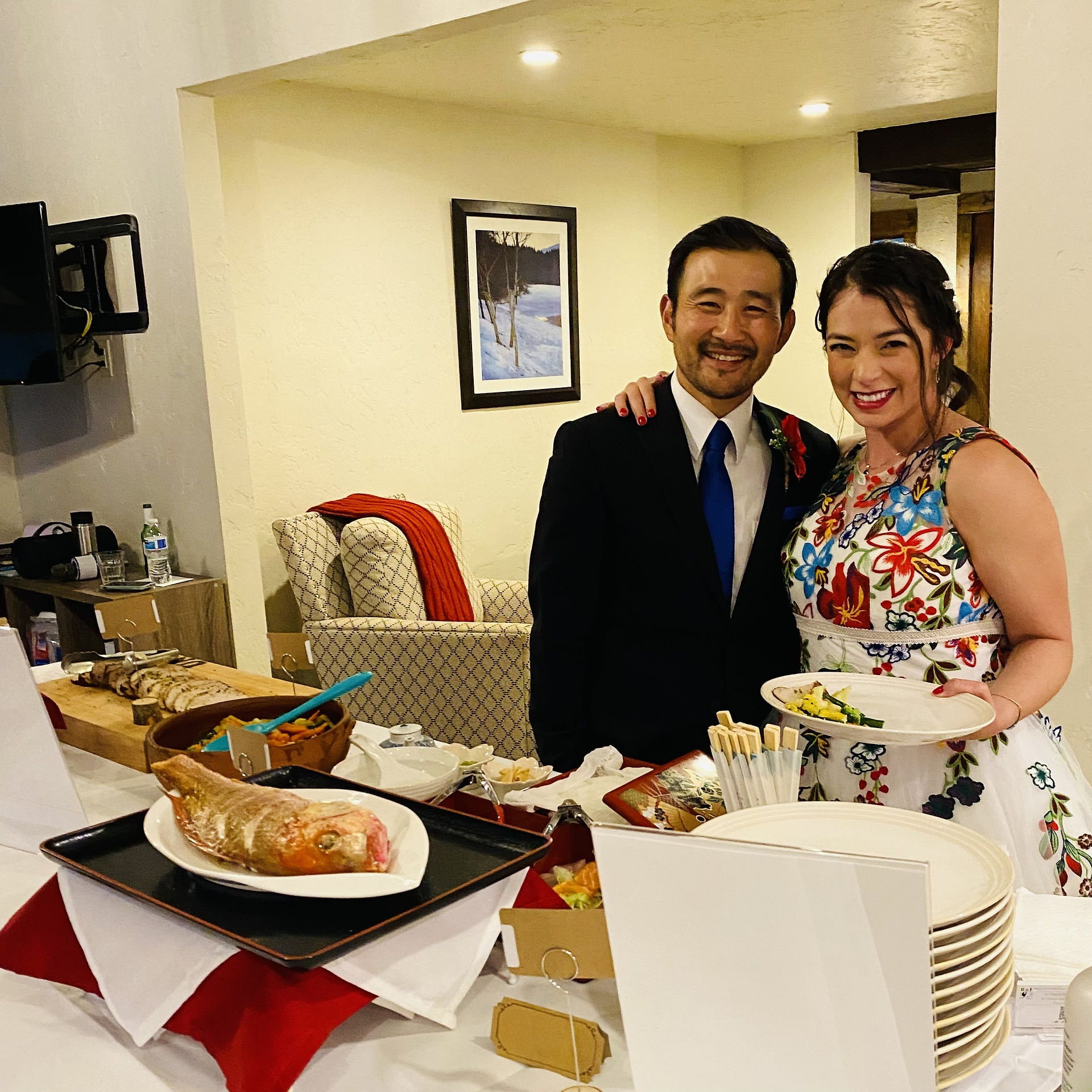 An Asian male with black suit and blue tie on the left, a Mexican heritage young woman next to him with beautiful wedding dress, full and colorful of flower embroidery stitches, holding a dinner plate over the elopement reception table, smiling at a camera