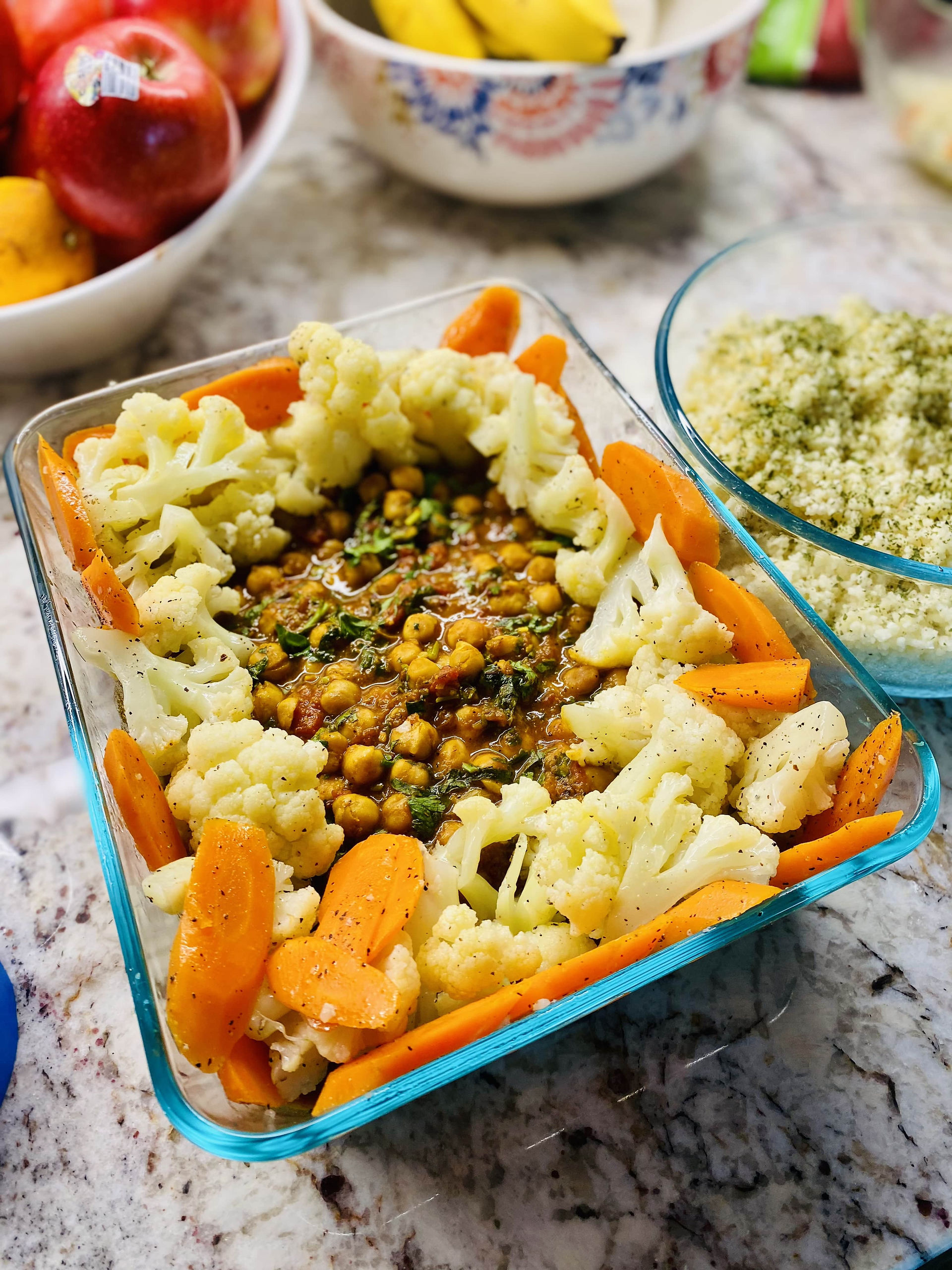 Chana Masala (Indian Vegetarian Curry with Chickpeas) in a center, blanched Cauliflower and Carrots around it in a rectangular glass container. Couscous Tabouleh in a smaller round glass container on the right. 