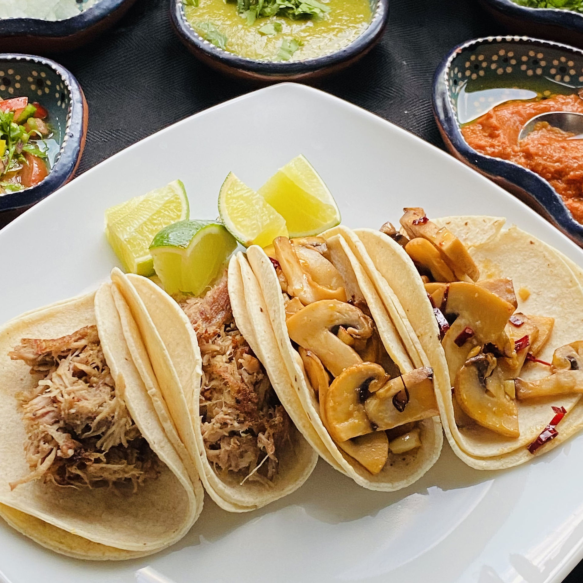 4 street tacos, from the left two carnitas in small corn tortillas, and two sauteed mushrooms and guajillo chili in two corn tortillas, a few cut limes on the side of the plate. There are some side dishes in small dishes with Mexican art on top of the photo. On the top left, pico de Gallo, green salsa with chopped cilantro on top, and red salsa with a spoon in on the right. 