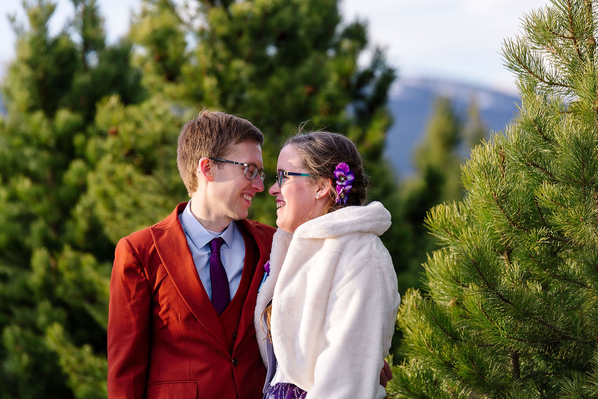 The wedding couple smiling at each other and stnading side by side, man in red suite and blue button up shirt, woman in white coat, both with rbown hair and glasses. Green trees in the background.