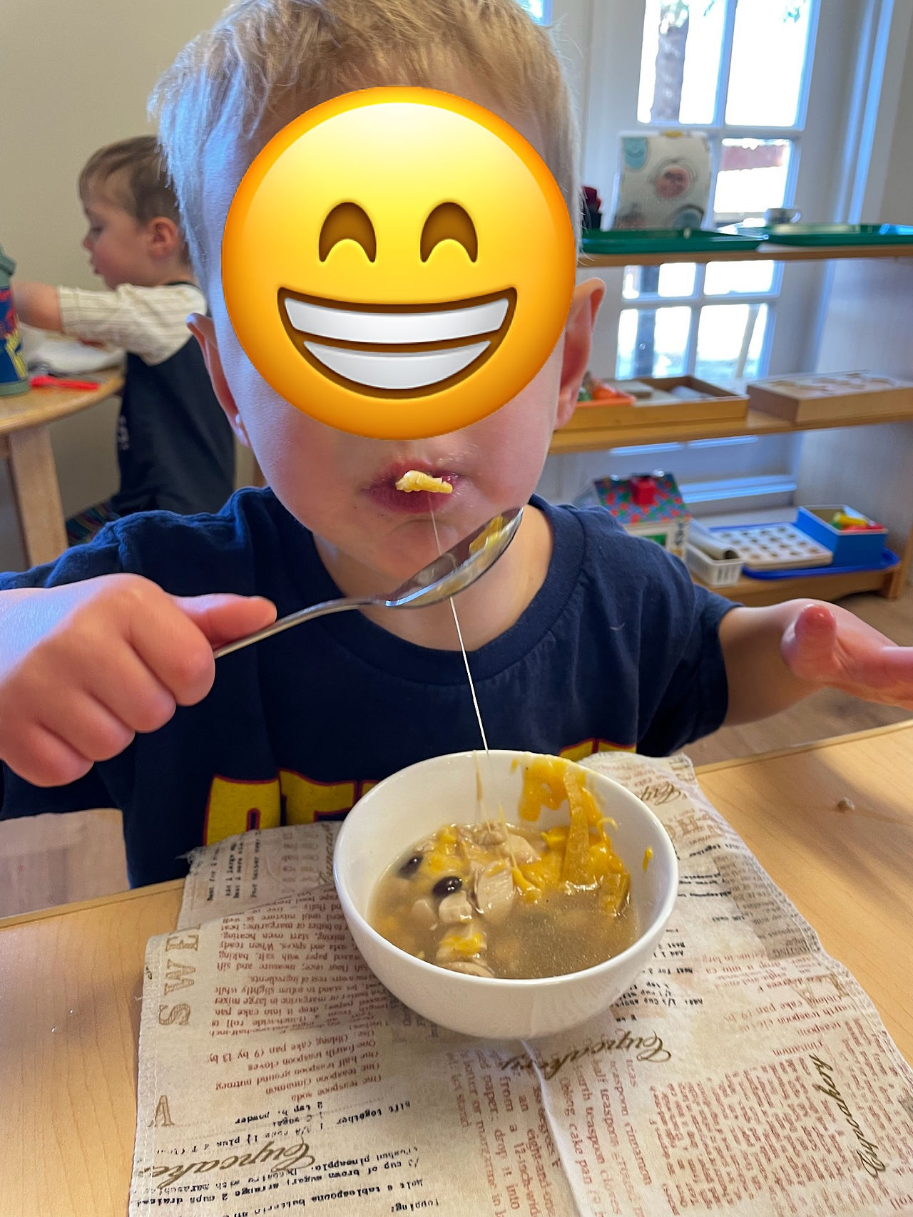 A 3 year old boy (face hidden by a sticker) holding a spoon with a string of melting cheese in between his soup bowl and his mouth
