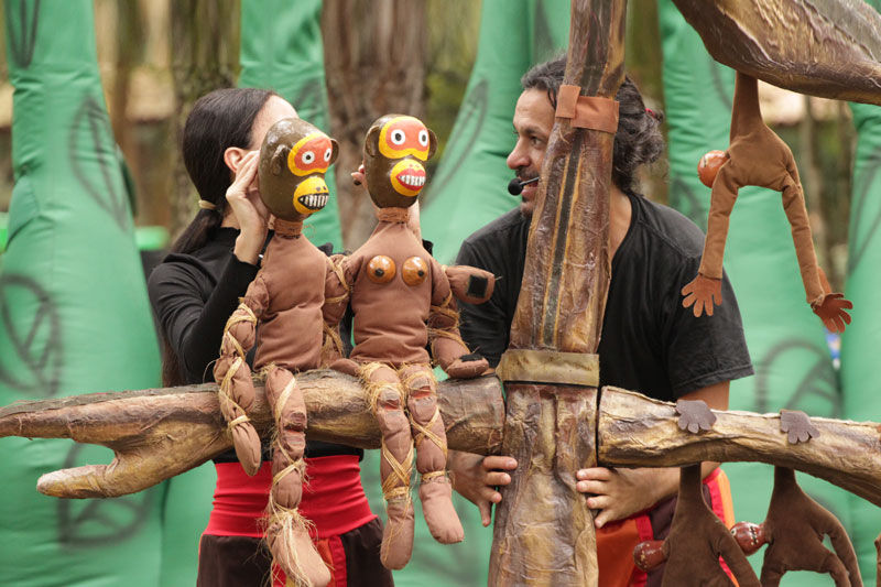 A imagem mostra dois atores, um homem e uma mulher segurando duas marionetes de macacos marrons, que estão sentados em um tronco de árvore