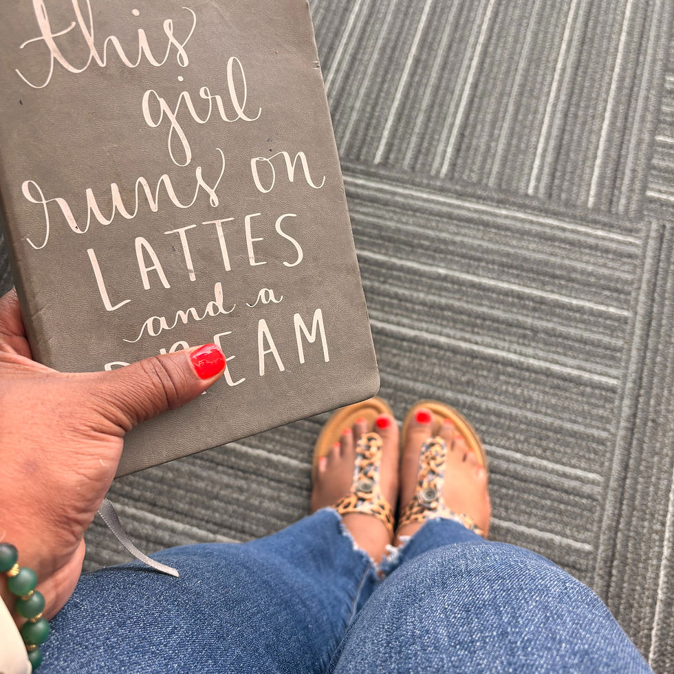 Hand holding notebook with text "This girl runs on lattes and a dream." Person in jeans with red toenails and sandals on gray striped floor.