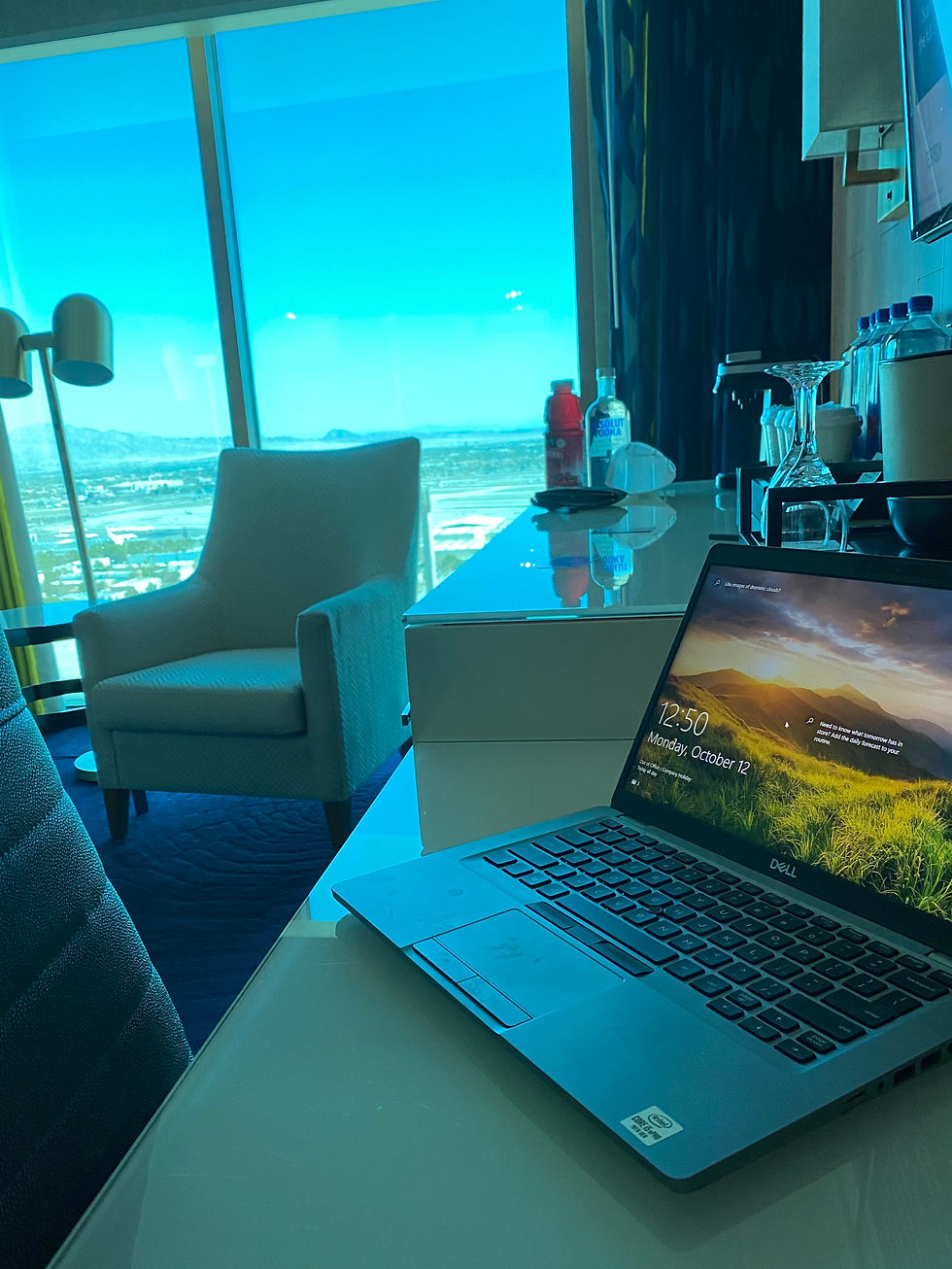Laptop on a desk in a hotel room with a mountain view. Bright blue sky, chair, lamp, and water bottles visible. Screen reads Monday, October 12.