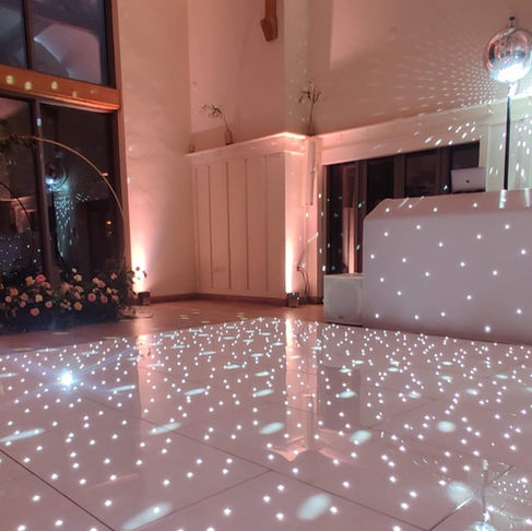 A beautifully decorated wedding venue with a shimmering dance floor lit up by a disco ball, creating an enchanting atmosphere. An elegant floral arch stands to one side, adding a touch of romantic charm to the celebration.