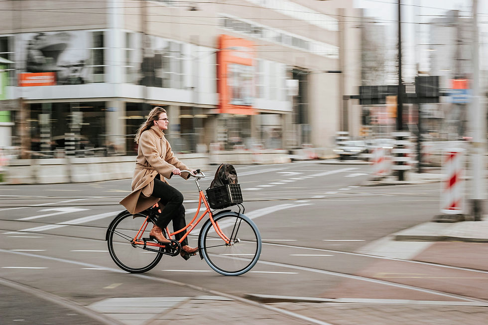 A woman wearing a tan coat and brown boots is riding an orange bicycle with a black basket on a city street. The background is blurred, showing motion.